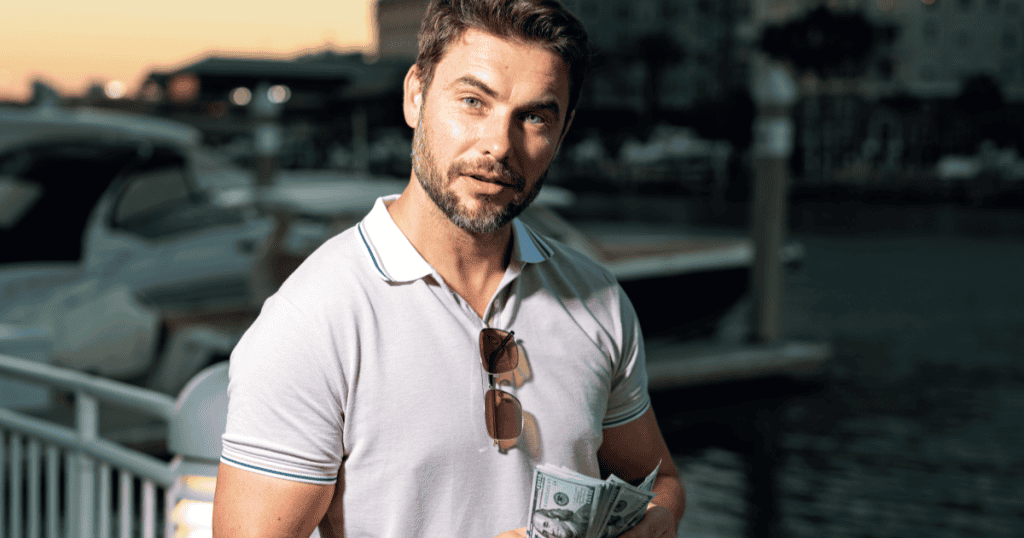 A well-dressed man casually holding cash by a marina, illustrating how a narcissistic man uses money as a tool of power and control to assert superiority over women.