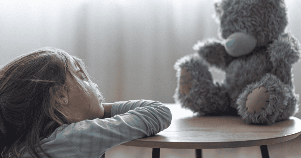 A young girl rests her head on her arms on a table across from a fluffy teddy bear, reflecting the quiet resilience of learning to nurture and protect your own inner vulnerabilities.