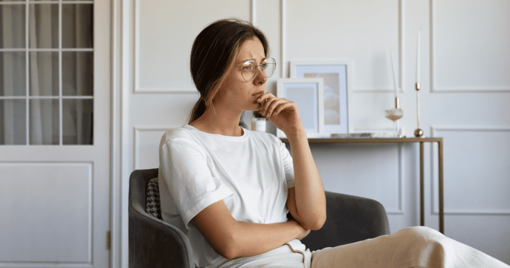 A woman wearing glasses sits in an armchair with a pensive expression, capturing the internal struggle of unravelling deeply ingrained self-doubt.