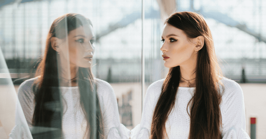 A woman in a white dress stands poised as she looks into her reflection on a glass wall, illustrating the unshakeable resolve required to stay centered when guilt is weaponized.