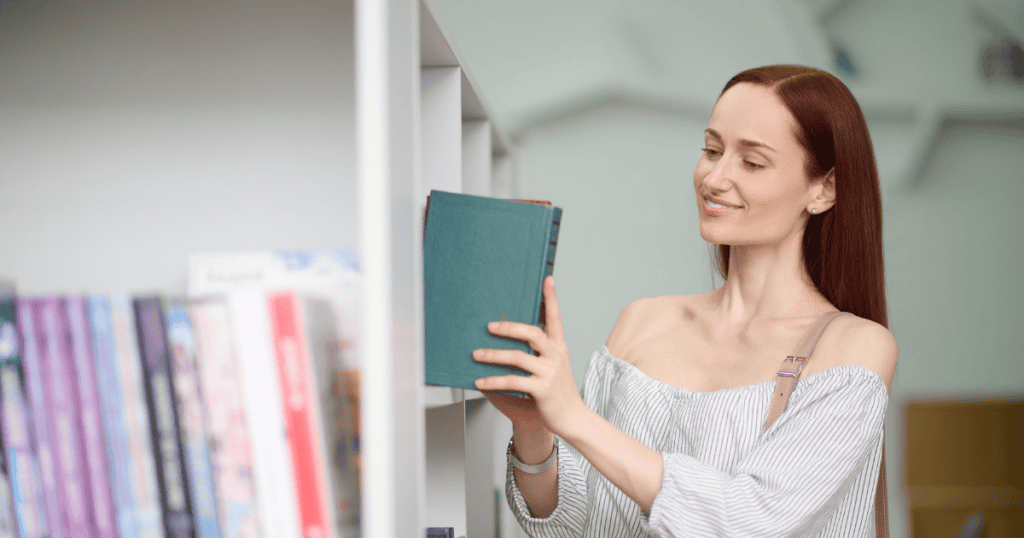 A woman smiles as she pulls a green book from a white library bookshelf, reflecting the clarity and empowerment found when finally understanding the patterns behind confusing communication.