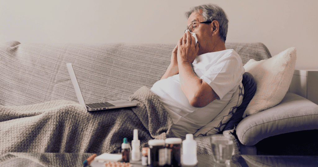 An older man lies under a blanket on a couch blowing his nose surrounded by medicine bottles and a laptop, highlighting how exaggerated helplessness is deployed to regain leverage.