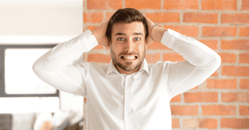 A man in a white dress shirt clutches both hands to the back of his head with a wide-eyed, anxious grimace against a brick wall, capturing the acute overwhelm and panic that strikes when reality finally catches up.