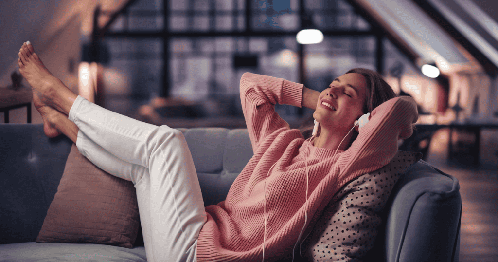 A woman in a pink sweater reclines comfortably on a sofa listening to music with her eyes closed and arms behind her head, capturing the pure comfort of resting completely unburdened.