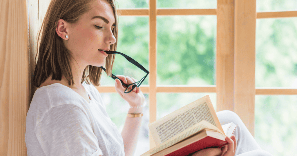 A woman thoughtfully holds the temple tip of her glasses to her mouth while reading an open book by a sunlit window, capturing the quiet insight and self-assurance gained from understanding relational dynamics.