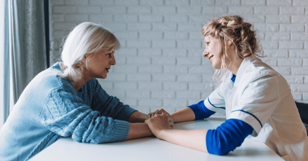 A woman holds hands across a table with a senior woman in a blue sweater, reflecting the calm, grounded reassurance of professional, compassionate care.