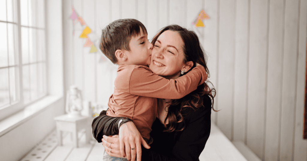 A young boy happily kisses his smiling mother on the cheek in a bright room decorated with bunting, showcasing the effortless warmth and joy found in a loving parent-child bond.