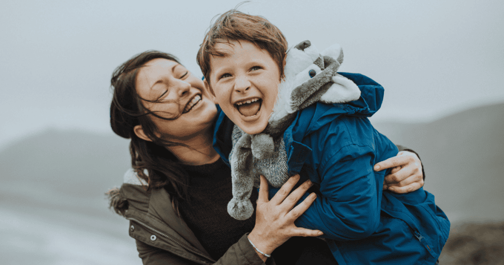 A mother and child laugh joyfully outdoors in the breeze while hugging a stuffed toy, embodying the carefree happiness and deep connection of unconditional love.