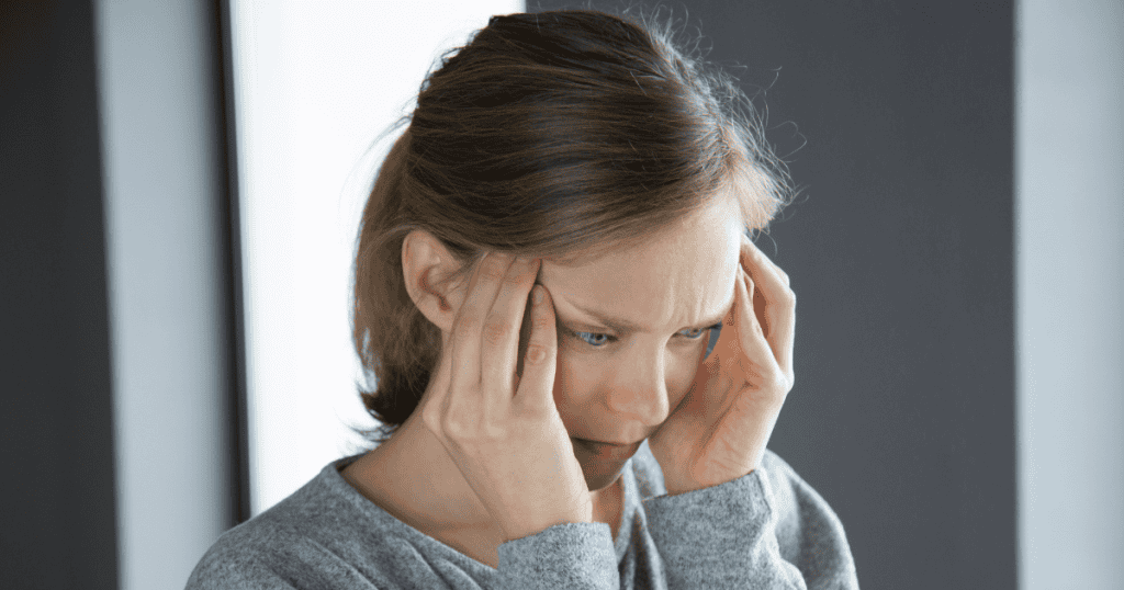 A distressed woman presses both hands against her temples with a downcast gaze, capturing the acute disorientation and self-doubt caused by selective amnesia tactics.
