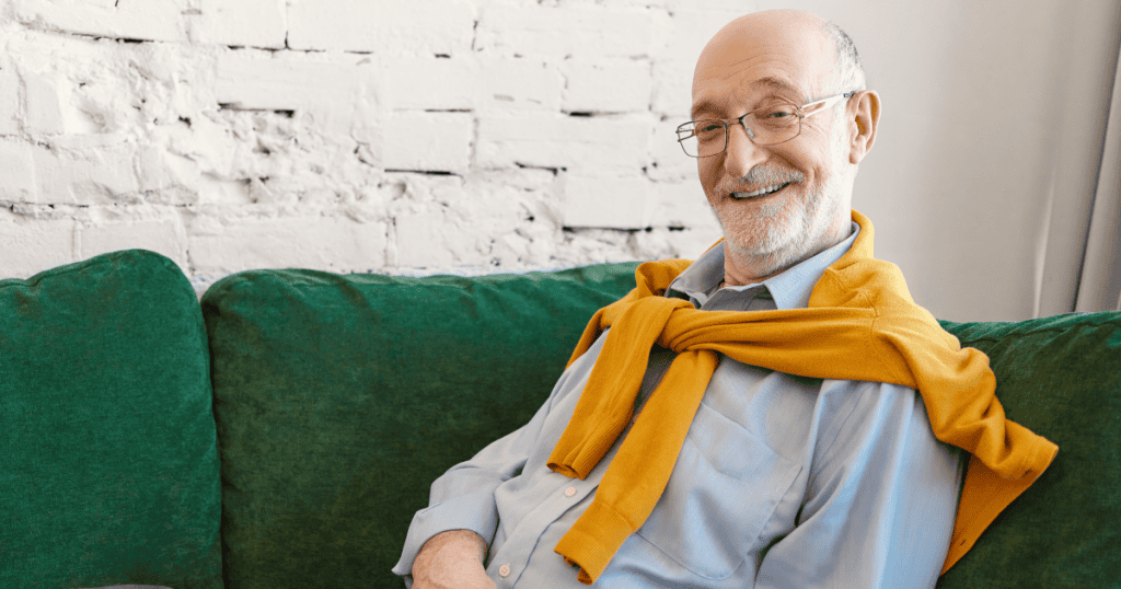 An older man in glasses smiles broadly while relaxing on a green couch, illustrating the confident, self-assured charm that can mask harder behavioral patterns emerging later in life.