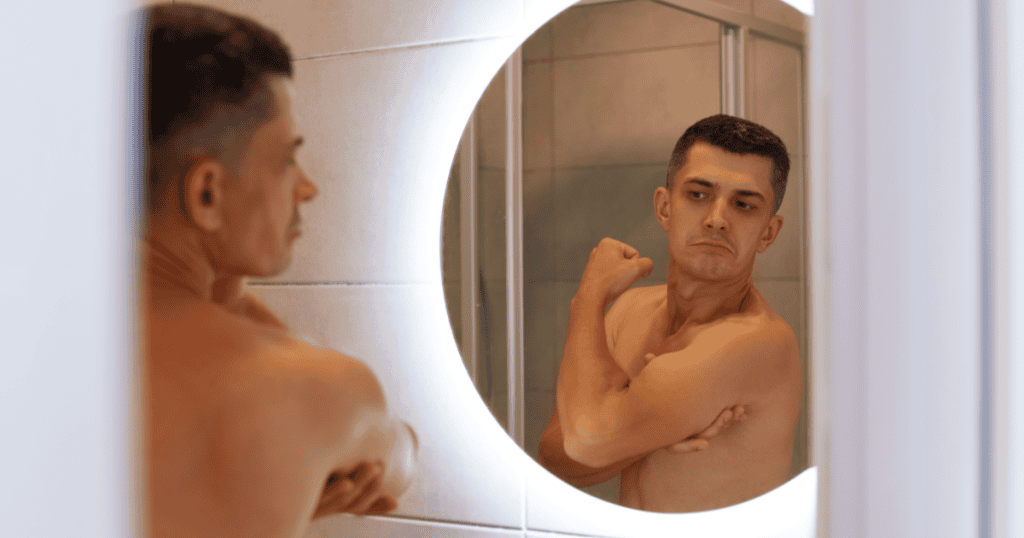 A shirtless man flexes his bicep while admiring himself in the bathroom mirror, embodying the self-admiration that can be an early warning sign of narcissism.