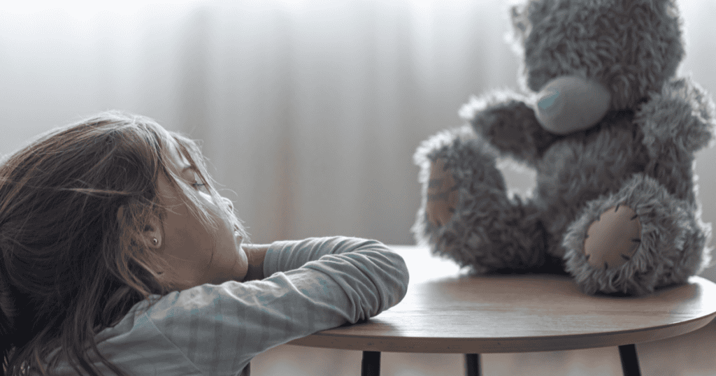 A small girl resting her head on a table, seeking comfort from a teddy bear, capturing the quiet loneliness of a child whose emotional needs went unmet in a narcissistic household.