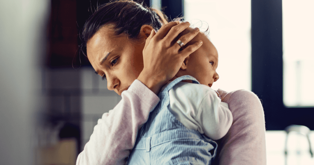 An exhausted mother holds her baby close over her shoulder with an anguished expression, capturing the deep ache of carrying early motherhood without genuine emotional support.