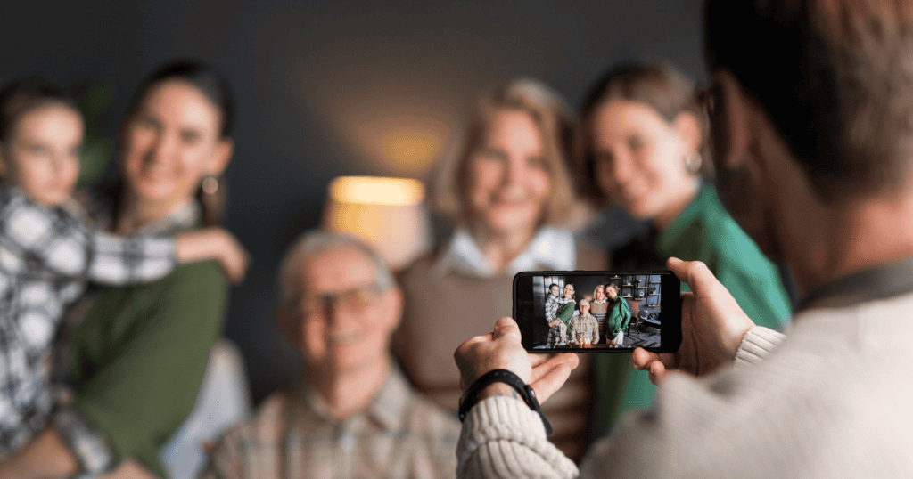 A person captures a group photo on a smartphone as a smiling family poses together in the background, illustrating the curated social presentation needed to protect external appearances.