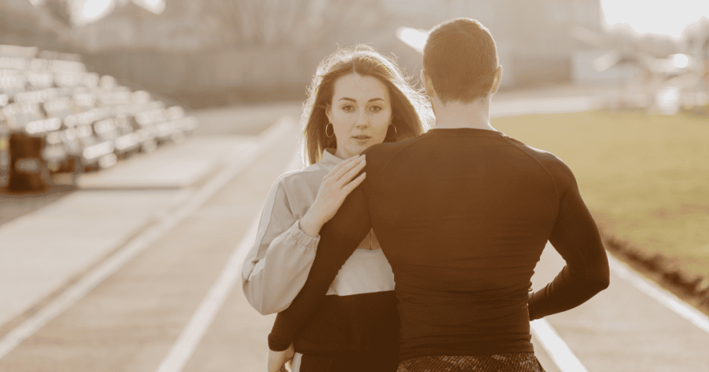 A woman places a hand on a man's shoulder while looking past him with a wary, distant expression, capturing the emotional pull that makes leaving take far longer than expected.
