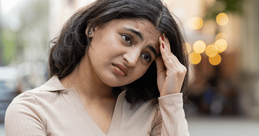 A woman presses fingers to her forehead with an anxious, conflicted frown outdoors, portraying the paralyzing doubt of having your independence constantly micro-managed.