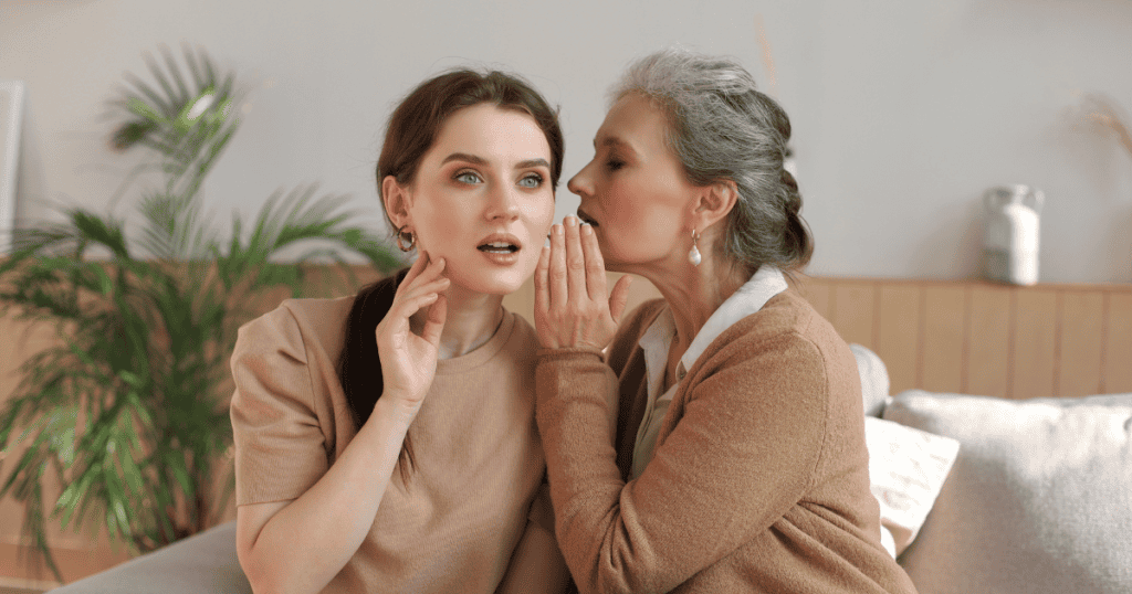 An older woman whispers closely into the ear of an uneasy young woman on a couch, portraying how boundary-setting often triggers covert, intrusive efforts to re-establish influence.