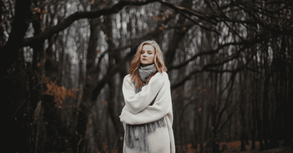 A young woman in a thick knit sweater stands with wrapped arms in a moody, bare forest, capturing the chilling quiet of confronting absolute solitude.