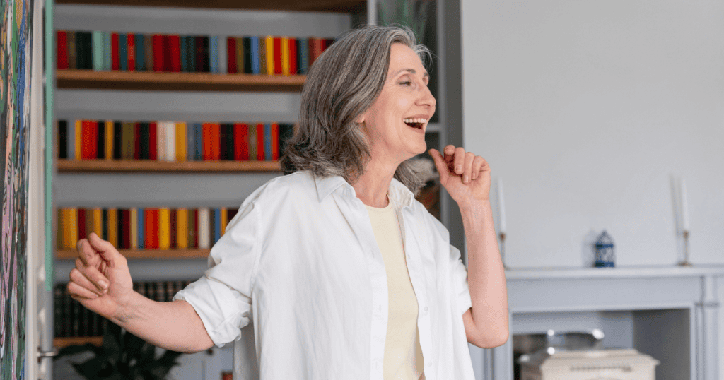 An older woman snaps her fingers and laughs happily in a bright room with colorful bookshelves, portraying an exaggerated display of unbothered cheer used to mask underlying tension.