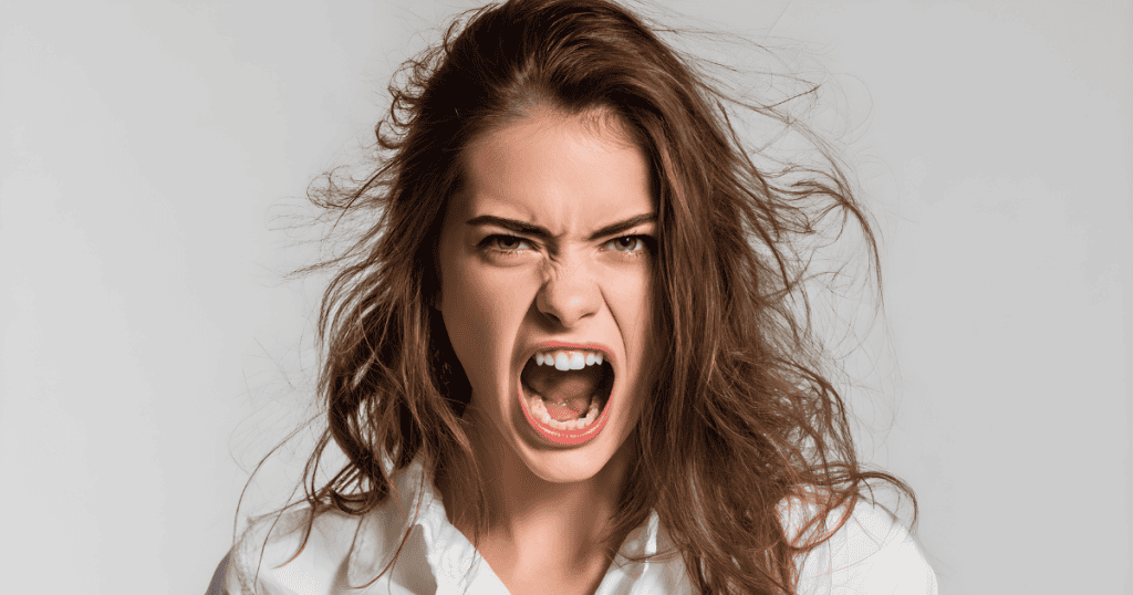 A young woman with windswept hair screams furiously with wide eyes against a neutral background, portraying an explosive release of built-up frustration and distress.