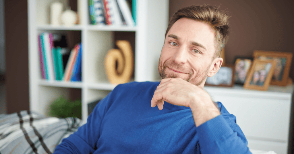 A man sitting comfortably at home with a smug, self-satisfied smirk, embodying the calm indifference a narcissist projects while their partner silently carries the weight of the abuse.