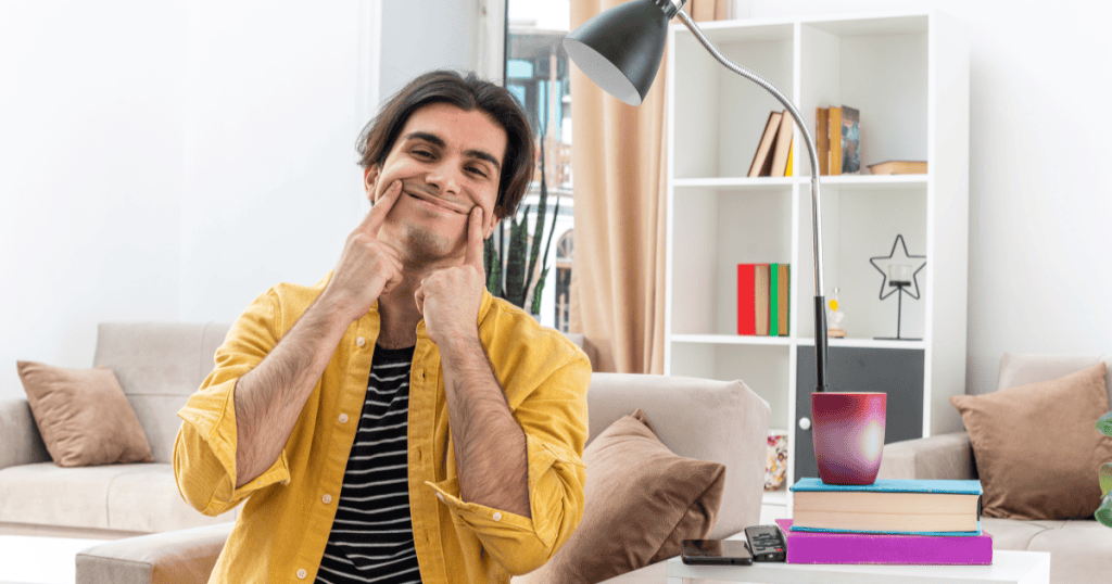 A man forces a smile by pushing up the corners of his mouth with his fingers, illustrating the manufactured happiness that may mask what's really underneath.