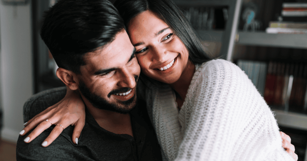 A smiling woman wraps her arms around a laughing man in a cozy, everyday moment, showing the kind of simple, genuine happiness with an "ordinary" partner that secretly humiliates the narcissist who was left behind.
