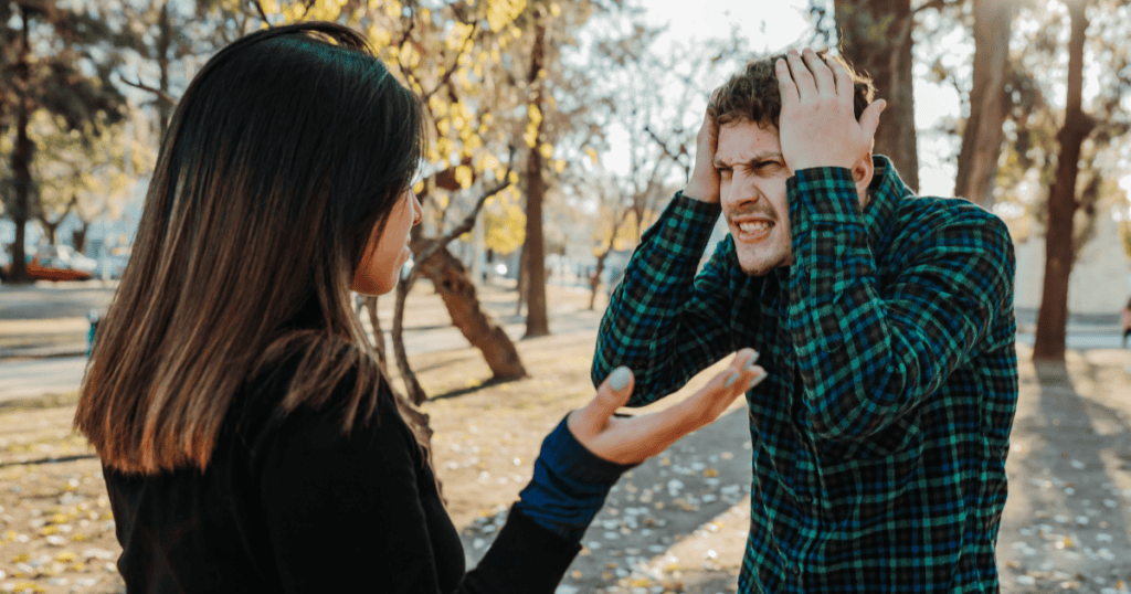 A man clutches his head in frustration while a woman gestures toward him during an outdoor argument, illustrating the tendency to blame others rather than take responsibility.