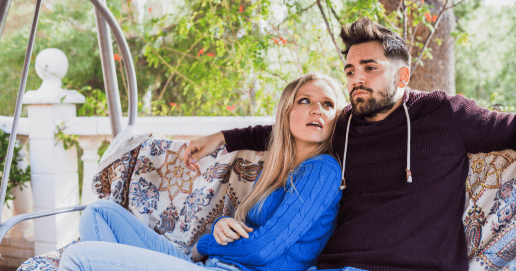 A couple sits close together on an outdoor swing, both looking away with unreadable, mismatched expressions, capturing the confusion of emotional inconsistency in a relationship.