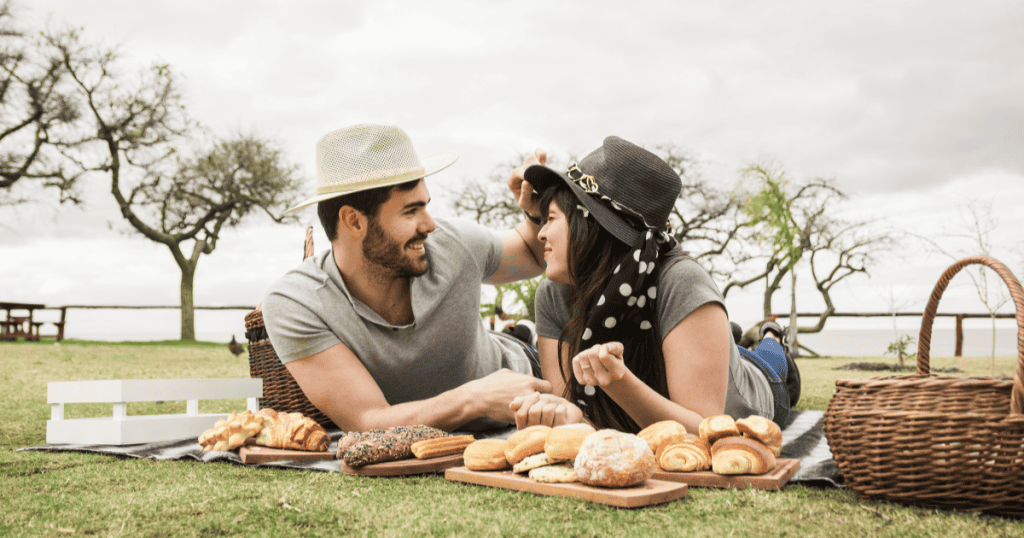 A couple lies close together sharing a laugh at a bread-filled picnic, illustrating the intense, all-consuming togetherness that can be an early sign of excessive attachment.