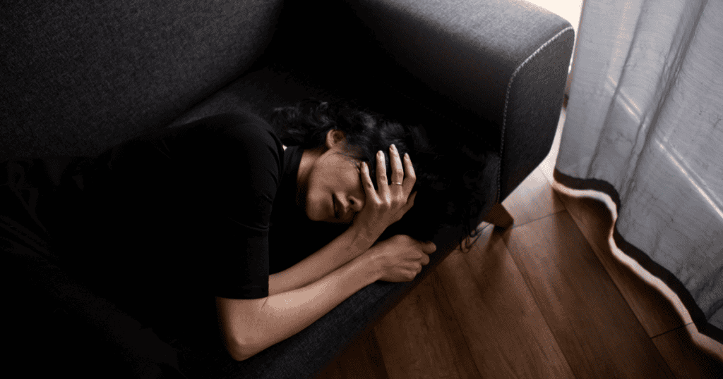 A woman lies curled on a dark sofa with her hand pressed over her eyes, capturing how fear can leave someone frozen in place with a narcissist long after logic says it's time to leave.