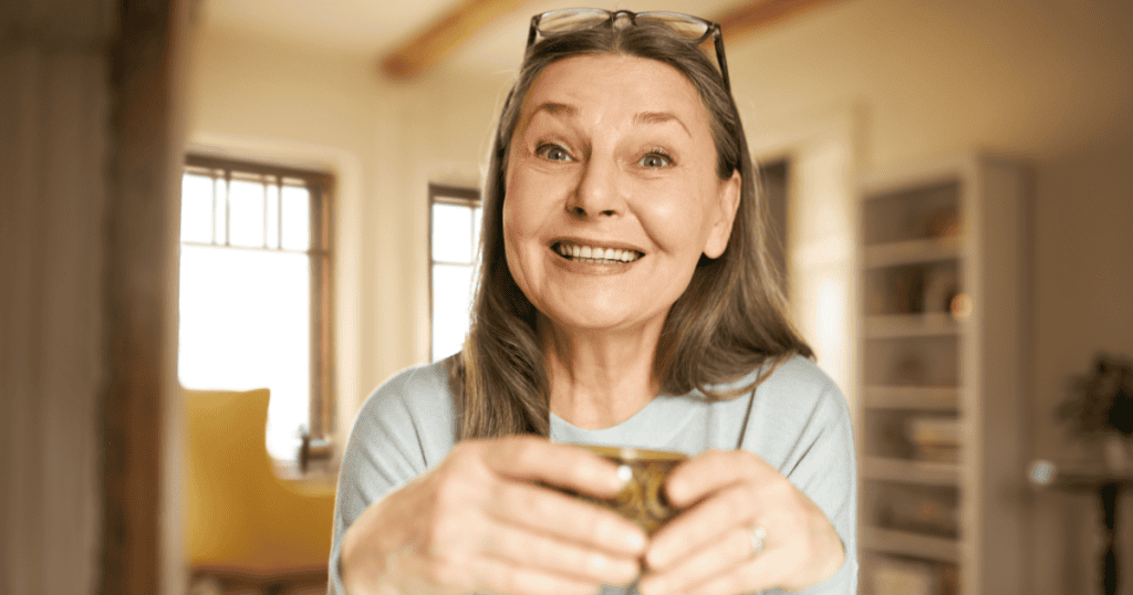 An older woman beaming with exaggerated enthusiasm as she extends a mug forward, capturing the convincing warmth a narcissist projects when making promises they have no intention of keeping.