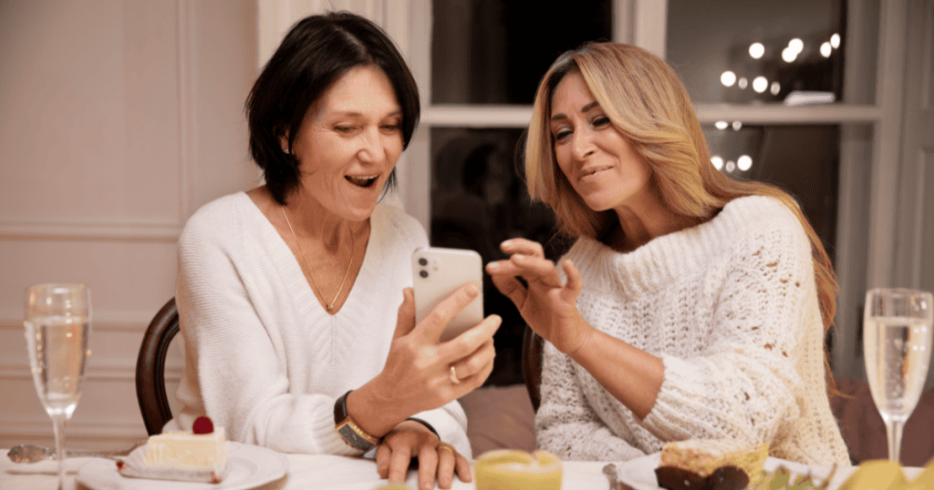 Two women eagerly sharing something on a phone over dinner, depicting how a narcissist casually exposes your private information to others as a way to gossip, control, or undermine you.