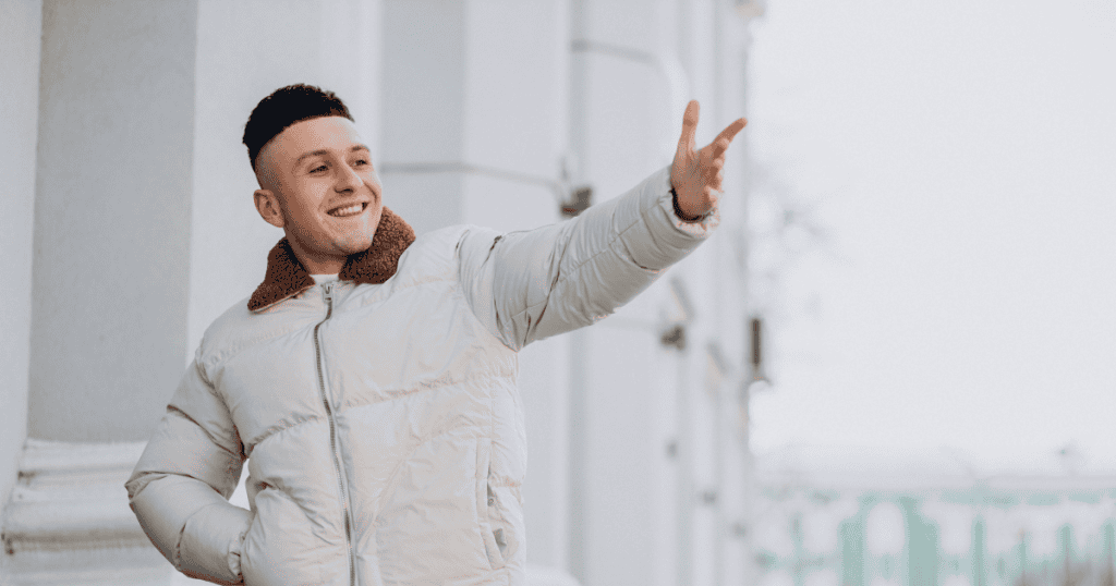 A man waves cheerfully with a bright smile while standing outdoors, embodying the outward confidence that serves as a cover rather than the truth underneath.