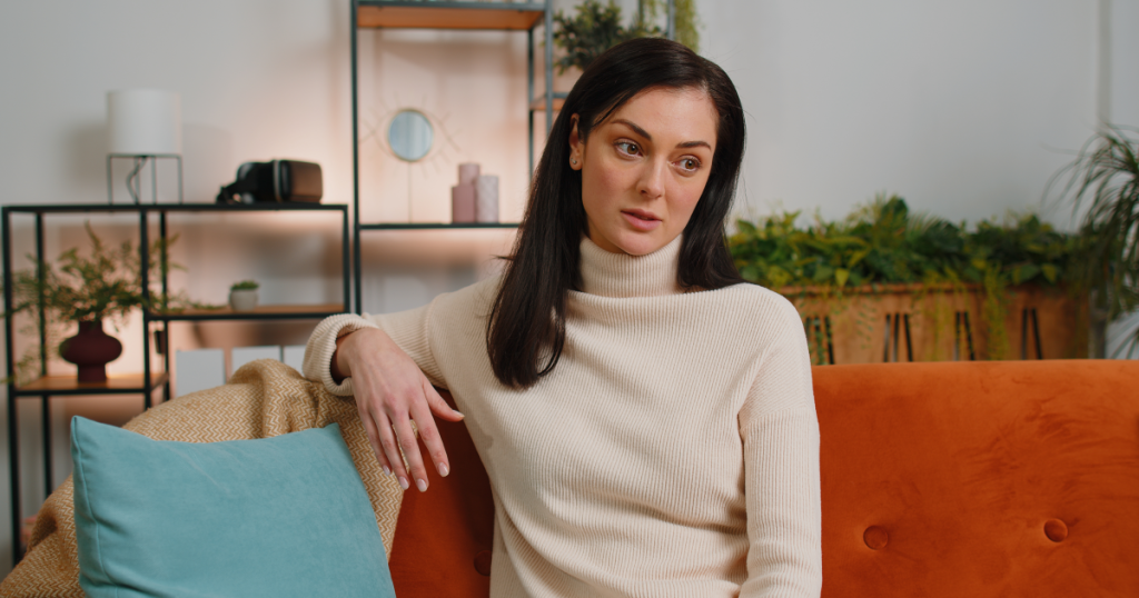 A woman sitting alone on a couch with a hollow, distant stare, capturing the quiet, numb anger of a survivor who has stopped screaming and started simply disconnecting.