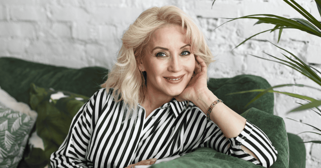 A woman poses with a warm, confident smile on a green couch, embodying the polished charm that can mask a deeper sense of loneliness underneath.