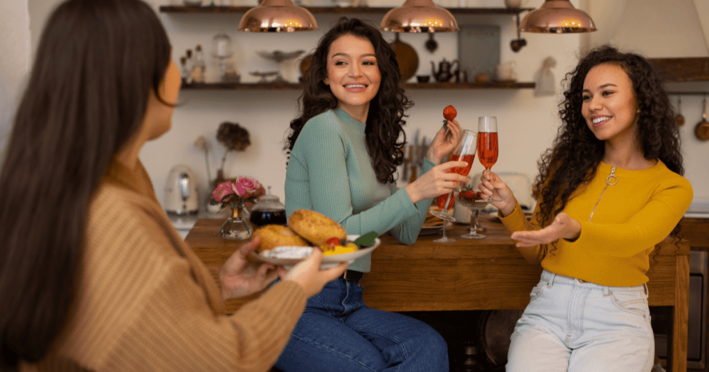 Two women raise drinks and eagerly welcome a friend bringing a plate of food to a stylish kitchen gathering, illustrating how a narcissist's warmth toward others is driven by what people can offer rather than who they truly are.