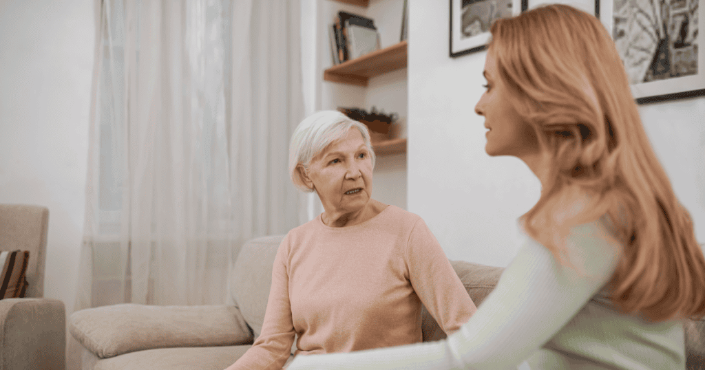 An older woman speaks earnestly on the couch while a younger woman listens intently, capturing the escalating urgency behind rewriting shared history.