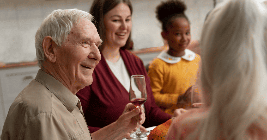 An elderly man smiles warmly while raising a glass of wine at a family gathering, evoking a nostalgic focus on cherished memories from the past.
