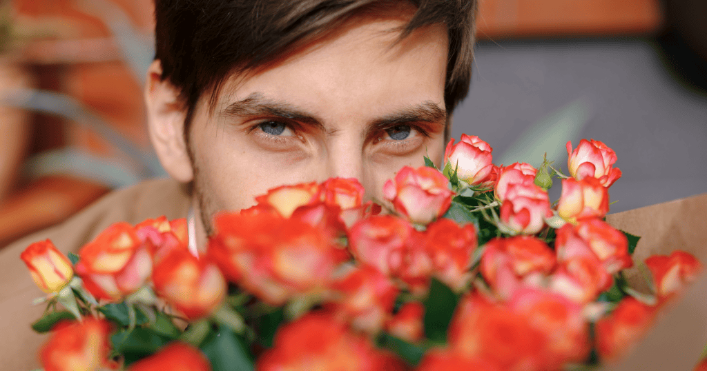 A man's eyes peer intently over a bouquet of roses, his gaze contrasting with the romantic gesture to hint at a too-good-to-be-true persona.