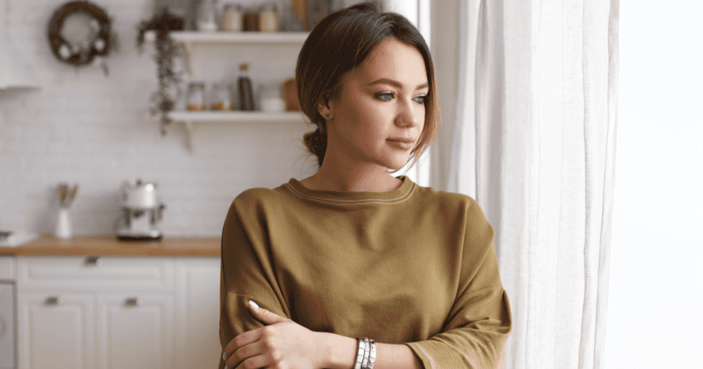 A woman stands by a sunlit window with her arms gently folded and a calm, reflective expression, suggesting the self-compassion that comes from understanding why leaving a narcissist takes longer than it "should."