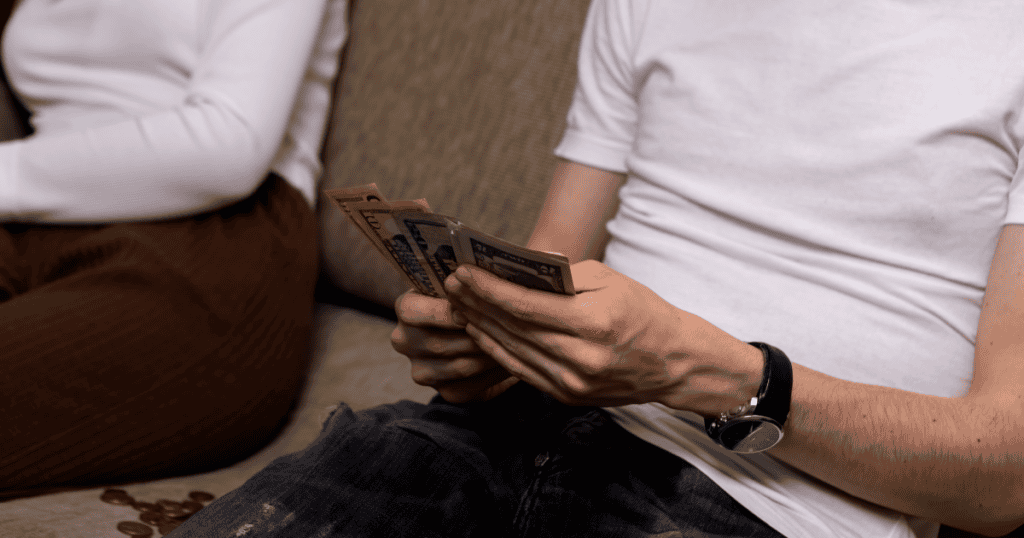A man counts cash on the couch while a woman sits with arms crossed nearby, illustrating how money can be wielded to define worth and reinforce a sense of position in the relationship.