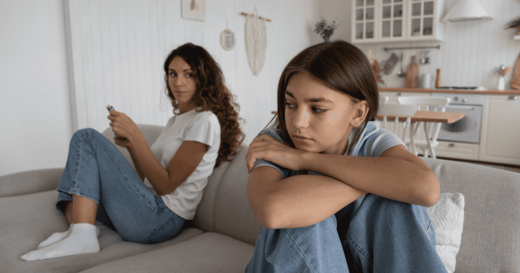 A young woman curls up hugging her knees with a withdrawn expression while another woman looks on holding her phone nearby, capturing the emotional distance that makes real intimacy feel out of reach.