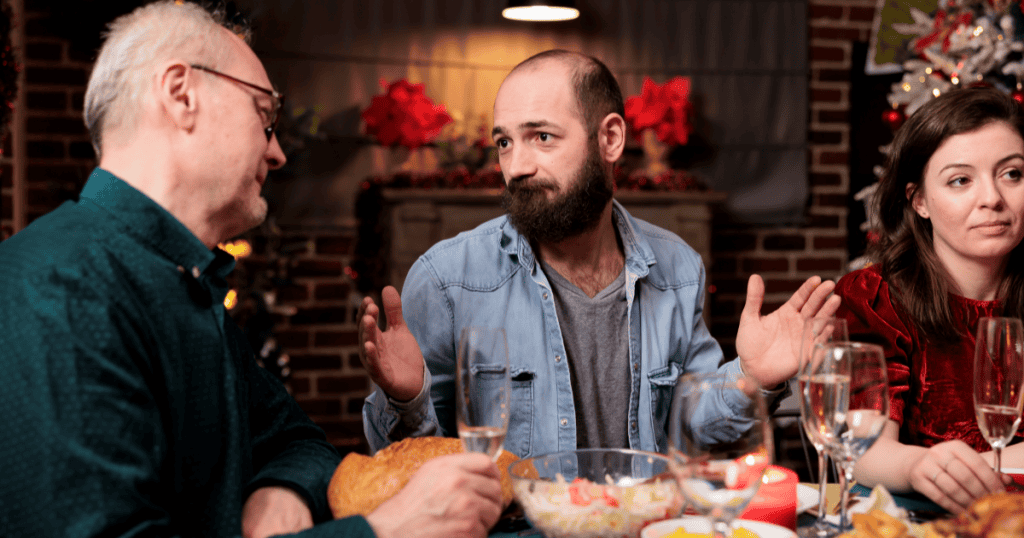 A man gestures defensively while explaining himself at a festive holiday dinner table, illustrating how self-preservation drives a narcissist to deflect blame even among family.