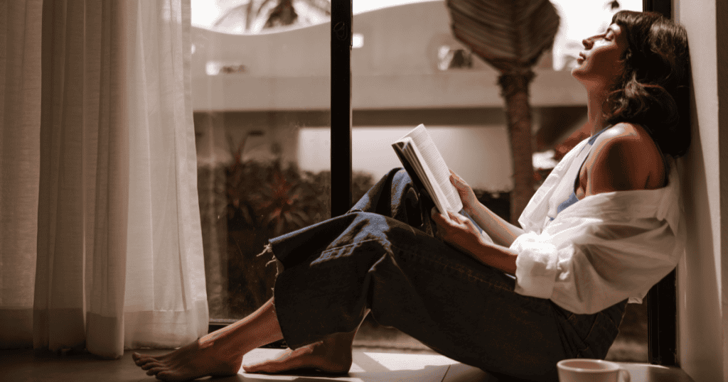 A woman leans back against a sunlit window with her eyes closed and a book in hand, embodying the sense of peace and release that comes with new understanding.