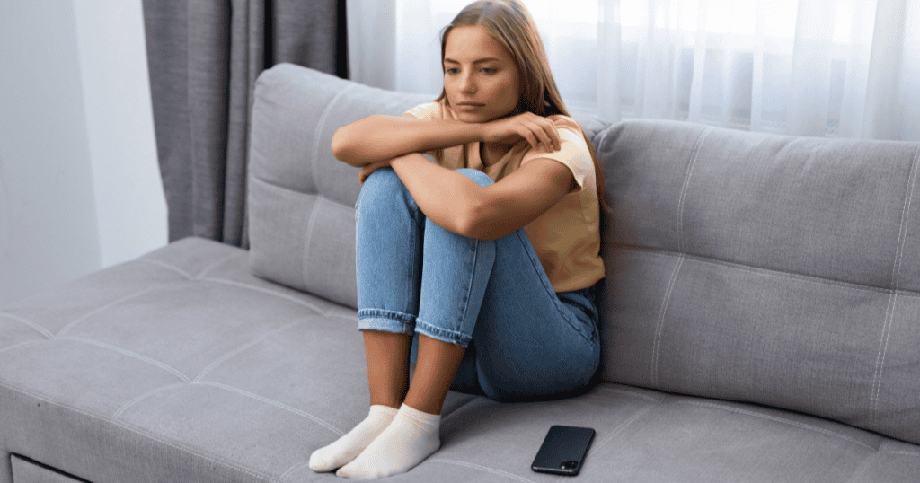 A young woman curled up alone on a couch with her phone untouched beside her, reflecting the deep isolation that follows those raised by a narcissist who made connection feel unsafe.