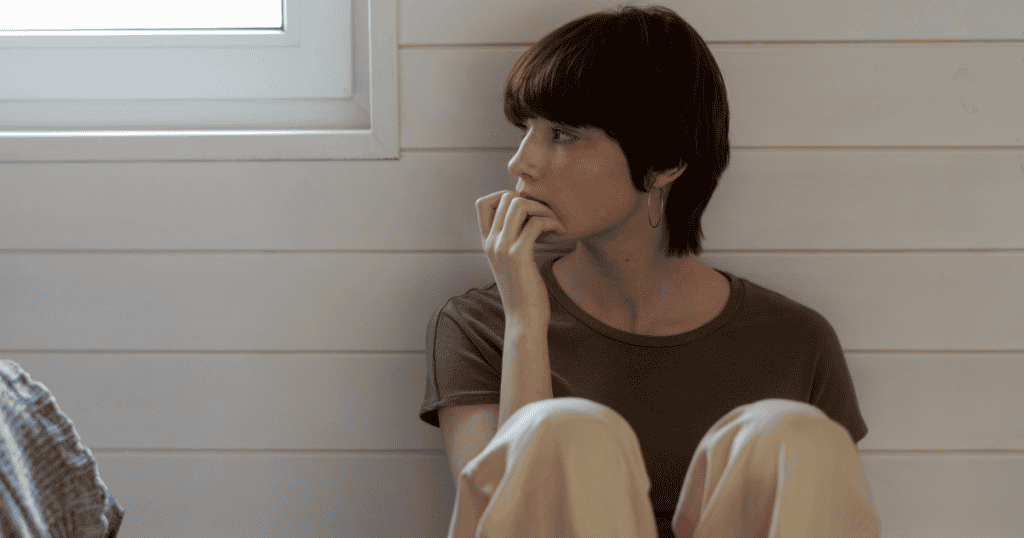 A woman sitting tensely on the floor with her hand to her mouth and a wary gaze, embodying the chronic, sourceless anxiety wired into someone raised in a narcissistic home.