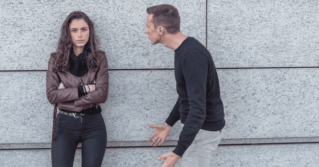 A woman with crossed arms stands guarded against a stone wall while a man leans toward her gesturing insistently, illustrating how early conditioning to doubt yourself can make a narcissist's pressure feel familiar and harder to walk away from.