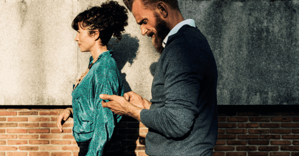 A woman walks away with a calm, distant expression while a bearded man stays absorbed in his phone behind her, reflecting how a trauma bond can keep the nervous system tethered to a narcissistic partner even as someone tries to leave.