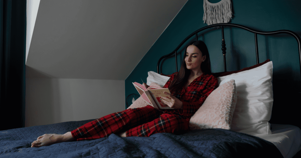 A woman in plaid pajamas relaxes on her bed smiling over a book, conveying the peace and ease that can follow leaving a narcissist, no matter how long the journey to freedom took.